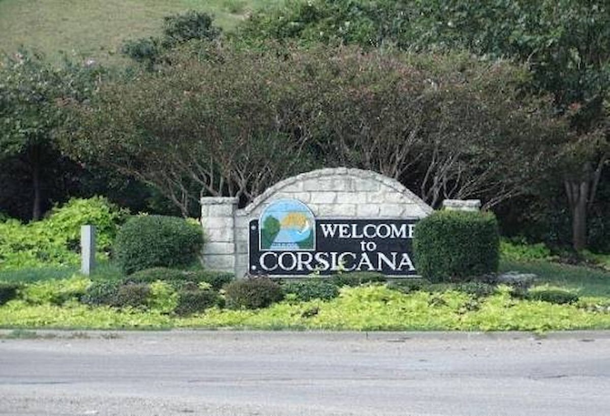 Stone welcome sign reading "Welcome to Corsicana" surrounded by green landscaping and trees.